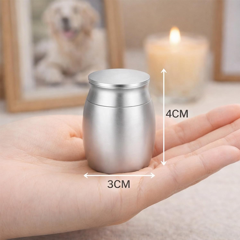 A small metallic silver cremation urn or keepsake jar is shown resting on an open hand, with overlaid dimension arrows and text indicating 3 cm width and 4 cm height. The background is softly blurred with a framed pet photo and a lit candle.