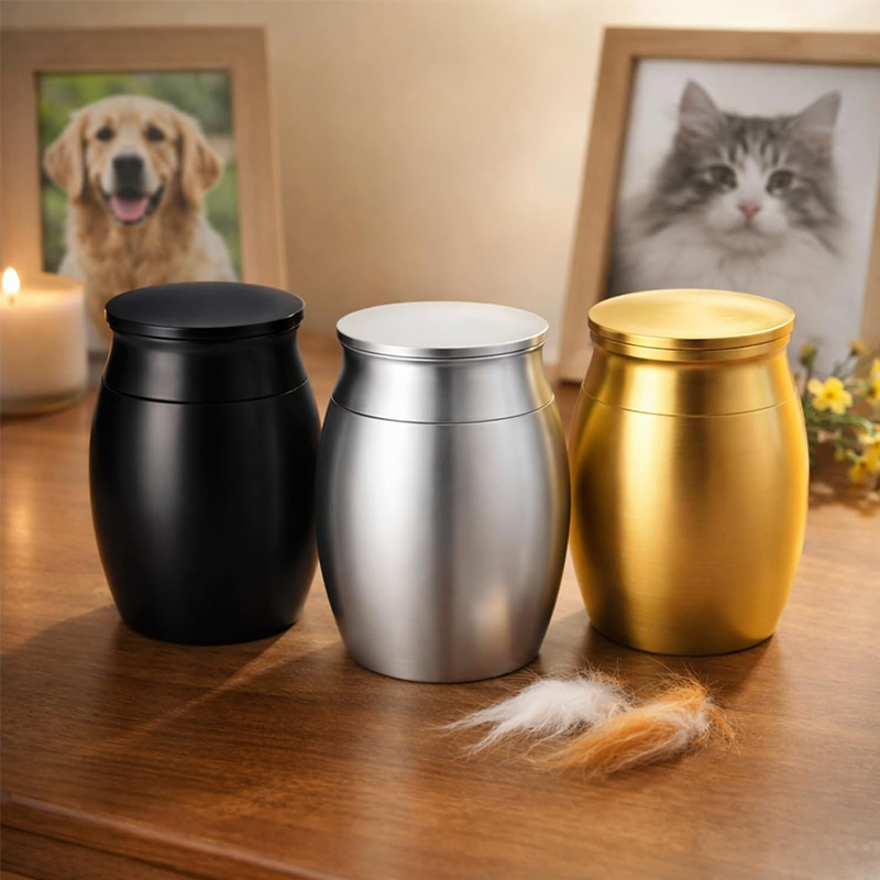 Three small metal urns with lids in black, silver, and gold, displayed on a wooden table with pet memorial props including a dog and cat photo frame, a candle, and a tuft of fur.